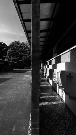 A black and white image of an outdoor facility with urinals lined up against a wall under an overhanging roof. The scene is split symmetrically by a vertical pillar, with sunlight casting strong shadows on the patterned floor.