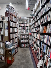 A narrow aisle in a bookstore is lined with tall wooden bookshelves filled with a variety of books. The shelves are organized with books of different colors and sizes, creating an orderly yet cozy atmosphere. Overhead fluorescent lights illuminate the space, casting a soft glow on the worn concrete floor. A small wooden table on the left holds pamphlets and a basket.