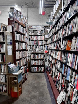 A narrow aisle in a bookstore is lined with tall wooden bookshelves filled with a variety of books. The shelves are organized with books of different colors and sizes, creating an orderly yet cozy atmosphere. Overhead fluorescent lights illuminate the space, casting a soft glow on the worn concrete floor. A small wooden table on the left holds pamphlets and a basket.