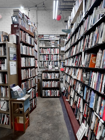 A narrow aisle in a bookstore is lined with tall wooden bookshelves filled with a variety of books. The shelves are organized with books of different colors and sizes, creating an orderly yet cozy atmosphere. Overhead fluorescent lights illuminate the space, casting a soft glow on the worn concrete floor. A small wooden table on the left holds pamphlets and a basket.