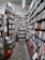 A narrow aisle in a bookstore is lined with tall wooden bookshelves filled with a variety of books. The shelves are organized with books of different colors and sizes, creating an orderly yet cozy atmosphere. Overhead fluorescent lights illuminate the space, casting a soft glow on the worn concrete floor. A small wooden table on the left holds pamphlets and a basket.