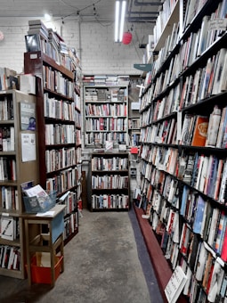 A narrow aisle in a bookstore is lined with tall wooden bookshelves filled with a variety of books. The shelves are organized with books of different colors and sizes, creating an orderly yet cozy atmosphere. Overhead fluorescent lights illuminate the space, casting a soft glow on the worn concrete floor. A small wooden table on the left holds pamphlets and a basket.
