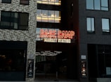 A neon sign with prominent red and yellow colors displays the words 'BASE CAMP MARKET STATION' on the facade of a building. The structure features modern architecture with dark and light toned brick patterns. Large windows are visible on either side of the sign.
