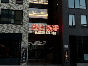 A neon sign with prominent red and yellow colors displays the words 'BASE CAMP MARKET STATION' on the facade of a building. The structure features modern architecture with dark and light toned brick patterns. Large windows are visible on either side of the sign.