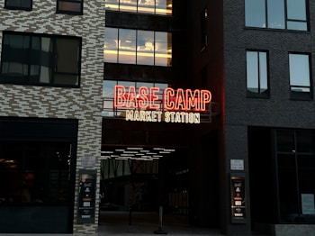 A neon sign with prominent red and yellow colors displays the words 'BASE CAMP MARKET STATION' on the facade of a building. The structure features modern architecture with dark and light toned brick patterns. Large windows are visible on either side of the sign.