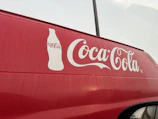 A red vehicle features a large white Coca-Cola logo prominently displayed on its side, accompanied by an outline of a Coca-Cola bottle. The background appears overcast, with faint reflections visible on the surface of the vehicle.