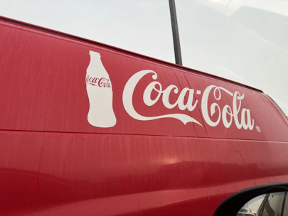 A red vehicle features a large white Coca-Cola logo prominently displayed on its side, accompanied by an outline of a Coca-Cola bottle. The background appears overcast, with faint reflections visible on the surface of the vehicle.