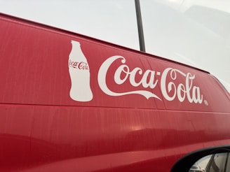 A red vehicle features a large white Coca-Cola logo prominently displayed on its side, accompanied by an outline of a Coca-Cola bottle. The background appears overcast, with faint reflections visible on the surface of the vehicle.