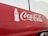 A red vehicle features a large white Coca-Cola logo prominently displayed on its side, accompanied by an outline of a Coca-Cola bottle. The background appears overcast, with faint reflections visible on the surface of the vehicle.