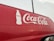 A red vehicle features a large white Coca-Cola logo prominently displayed on its side, accompanied by an outline of a Coca-Cola bottle. The background appears overcast, with faint reflections visible on the surface of the vehicle.