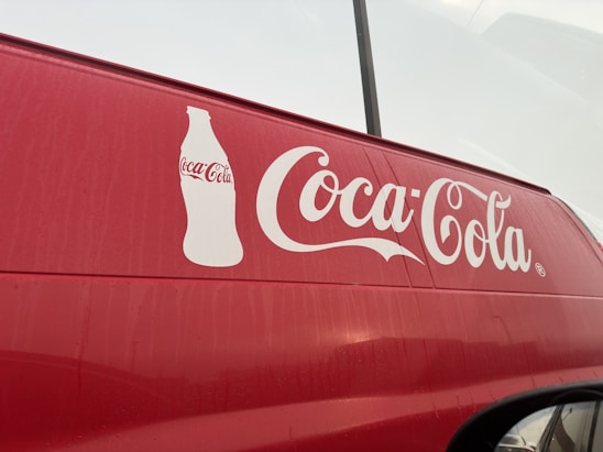 A red vehicle features a large white Coca-Cola logo prominently displayed on its side, accompanied by an outline of a Coca-Cola bottle. The background appears overcast, with faint reflections visible on the surface of the vehicle.