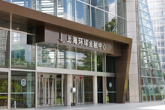 Entrance to a modern office building with large glass doors and windows, featuring the words 'Shanghai World Financial Center' in Chinese above the entrance. The reflection of surrounding skyscrapers is visible in the glass.