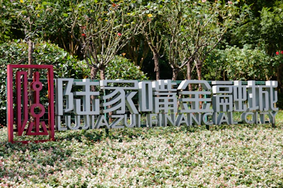 A sign with Chinese characters and the English text 'Lujiazui Financial City' is situated in a garden setting. The surrounding area is lush with greenery and leafy plants, and there is a stylized red frame on the left side of the text.