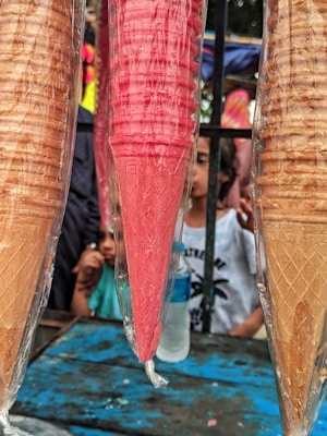 Three bundles of stacked ice cream cones, covered in plastic wrap. The center cone is red, while the two on either side are beige. The background includes blurred images of people, a metal fence, and colorful outdoor surroundings.
