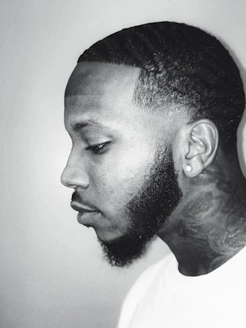 A monochrome portrait of a person in profile view. The focus is on the person's well-groomed hairstyle, facial hair, and tattooed neck.