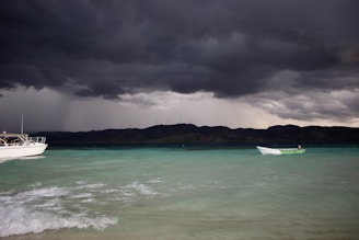 Dark storm clouds loom over a coastal scene where the sea is a mixture of turquoise and deep blue. Two boats are present, one larger and partially visible on the left and another smaller one in the distance, near the center. The background features low hills dotted with wind turbines.