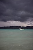 A small white boat floats on calm, turquoise waters under a dramatic sky filled with dark, stormy clouds. In the distance, a mountainous landscape is visible along with a line of wind turbines on the ridge.