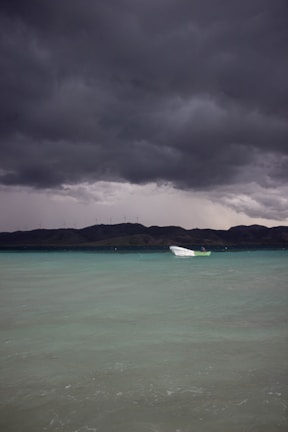 A small white boat floats on calm, turquoise waters under a dramatic sky filled with dark, stormy clouds. In the distance, a mountainous landscape is visible along with a line of wind turbines on the ridge.