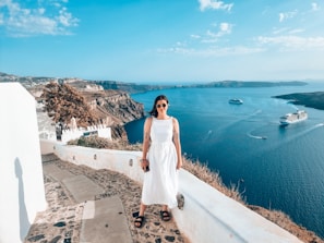 A person in a white dress stands on a scenic walkway overlooking a vast blue sea with cruise ships in the distance. Surrounding the pathway are white buildings and rocky cliffs typical of a Mediterranean landscape.