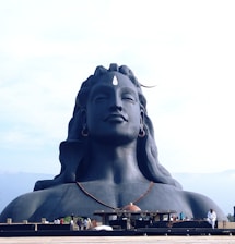 A large, serene statue depicting a figure with closed eyes and elongated earlobes wearing a crescent moon and a rudraksha necklace. The figure is made of a dark material, and it stands against a light blue sky background.