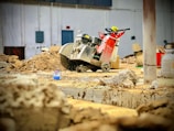 A construction site features a large industrial saw with a bright red and silver design, positioned among piles of dirt and debris. The background includes various building materials like boxes and containers, and a partially visible white wall with electrical panels.