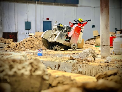 A construction site features a large industrial saw with a bright red and silver design, positioned among piles of dirt and debris. The background includes various building materials like boxes and containers, and a partially visible white wall with electrical panels.
