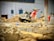 A construction site features a large industrial saw with a bright red and silver design, positioned among piles of dirt and debris. The background includes various building materials like boxes and containers, and a partially visible white wall with electrical panels.