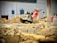 A construction site features a large industrial saw with a bright red and silver design, positioned among piles of dirt and debris. The background includes various building materials like boxes and containers, and a partially visible white wall with electrical panels.