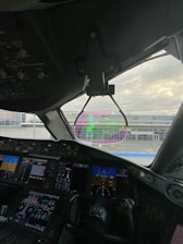 The interior of an aircraft cockpit features multiple digital displays and controls. A heads-up display is visible through the windshield, showing flight information with a view of the airport terminal and sky in the background.