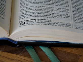 An open book lies on a wooden surface, capturing a close-up view of a page with text in a serif font. The visible text includes religious content and a highlighted bold section with the number 13. Green ribbon bookmarks are peeking out from the bottom of the book.