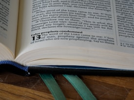An open book lies on a wooden surface, capturing a close-up view of a page with text in a serif font. The visible text includes religious content and a highlighted bold section with the number 13. Green ribbon bookmarks are peeking out from the bottom of the book.