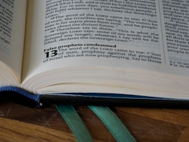 An open book lies on a wooden surface, capturing a close-up view of a page with text in a serif font. The visible text includes religious content and a highlighted bold section with the number 13. Green ribbon bookmarks are peeking out from the bottom of the book.