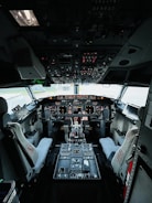 The cockpit of an aircraft filled with numerous controls and instruments. Two pilot seats are positioned in front of a dashboard equipped with multiple screens and switches. The overhead panel is densely packed with various knobs and lights. The overall setup suggests a highly technical and functional environment.