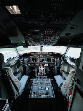 The cockpit of an aircraft filled with numerous controls and instruments. Two pilot seats are positioned in front of a dashboard equipped with multiple screens and switches. The overhead panel is densely packed with various knobs and lights. The overall setup suggests a highly technical and functional environment.