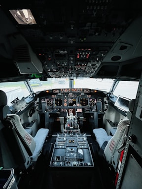 The cockpit of an aircraft filled with numerous controls and instruments. Two pilot seats are positioned in front of a dashboard equipped with multiple screens and switches. The overhead panel is densely packed with various knobs and lights. The overall setup suggests a highly technical and functional environment.