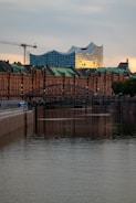 Modern architecture with a wave-like roof design stands prominently against a backdrop of traditional brick buildings. The scene is set alongside a calm waterway, with a bridge and a construction crane visible, adding an urban touch. The evening light casts a warm glow, highlighting the contrasting styles of the architecture.