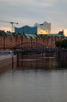 Modern architecture with a wave-like roof design stands prominently against a backdrop of traditional brick buildings. The scene is set alongside a calm waterway, with a bridge and a construction crane visible, adding an urban touch. The evening light casts a warm glow, highlighting the contrasting styles of the architecture.
