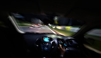 A view from inside a car looking out towards the road at night, with motion blur creating streaks of lights from surrounding streetlights and vehicles. The dashboard is illuminated with lights, and a person is visible holding the steering wheel.