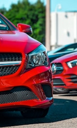 Close-up view of the front end of a red luxury car, prominently displaying the headlight and front grille. The background includes another similar car slightly out of focus, hinting at an outdoor setting, possibly a parking lot or dealership with blurred trees and a building in the backdrop.