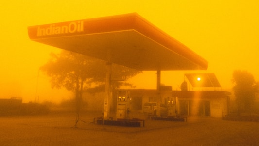 A gas station with the logo 'IndianOil' appears amid an intense yellow-orange haze, likely caused by a sandstorm or heavy pollution. The sky and surroundings are enveloped in this thick hue, significantly reducing visibility. The gas pumps are visible under the shelter, and there's a building in the background with a solar panel on its roof.