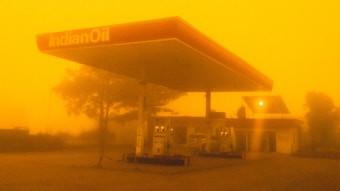 A gas station with the logo 'IndianOil' appears amid an intense yellow-orange haze, likely caused by a sandstorm or heavy pollution. The sky and surroundings are enveloped in this thick hue, significantly reducing visibility. The gas pumps are visible under the shelter, and there's a building in the background with a solar panel on its roof.