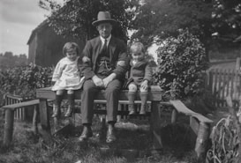 A man in a suit and hat sits on a wooden platform outdoors, surrounded by two young children. The man, positioned in the center, has his arms around both children. The child on the left wears a light-colored dress and boots, while the child on the right wears a dark coat and knee-high socks. They are in a rustic setting with a wooden fence and lush greenery in the background, including trees and bushes.