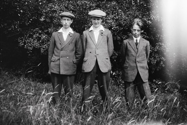 Three young boys, dressed in similar suits and caps, are standing in a grassy area with bushes in the background. The boys are arranged in a line, with two of them holding their hands behind their backs. The image is in black and white, giving it a vintage look.