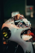 A vintage motorcycle with a white chassis, featuring a round yellow headlight. Various stickers adorn the top of the motorcycle, adding a colorful and personalized touch. The background is blurred, focusing attention on the motorcycle's details.