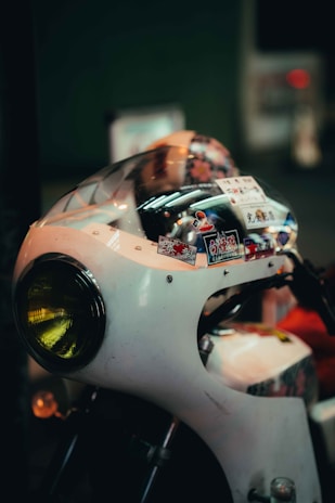A vintage motorcycle with a white chassis, featuring a round yellow headlight. Various stickers adorn the top of the motorcycle, adding a colorful and personalized touch. The background is blurred, focusing attention on the motorcycle's details.