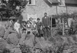 A vintage black and white photograph of a group of people, including both adults and children, sitting and standing on a stone wall in front of a rustic stone house. The individuals are dressed in period clothing, suggesting an early 20th-century setting. Some are seated, with one person holding an infant, while others stand beside them.