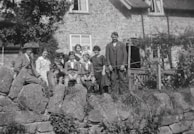 A vintage black and white photograph of a group of people, including both adults and children, sitting and standing on a stone wall in front of a rustic stone house. The individuals are dressed in period clothing, suggesting an early 20th-century setting. Some are seated, with one person holding an infant, while others stand beside them.