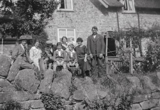 A vintage black and white photograph of a group of people, including both adults and children, sitting and standing on a stone wall in front of a rustic stone house. The individuals are dressed in period clothing, suggesting an early 20th-century setting. Some are seated, with one person holding an infant, while others stand beside them.