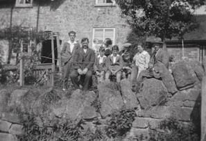 A group of people, including adults and children, is gathered outdoors in front of a stone wall and a house with a stone exterior. The people are dressed in early 20th-century clothing and are sitting and standing, with some looking at the camera. A tree and some plants are visible in the background.