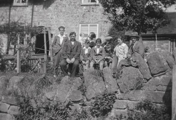 A group of people, including adults and children, is gathered outdoors in front of a stone wall and a house with a stone exterior. The people are dressed in early 20th-century clothing and are sitting and standing, with some looking at the camera. A tree and some plants are visible in the background.